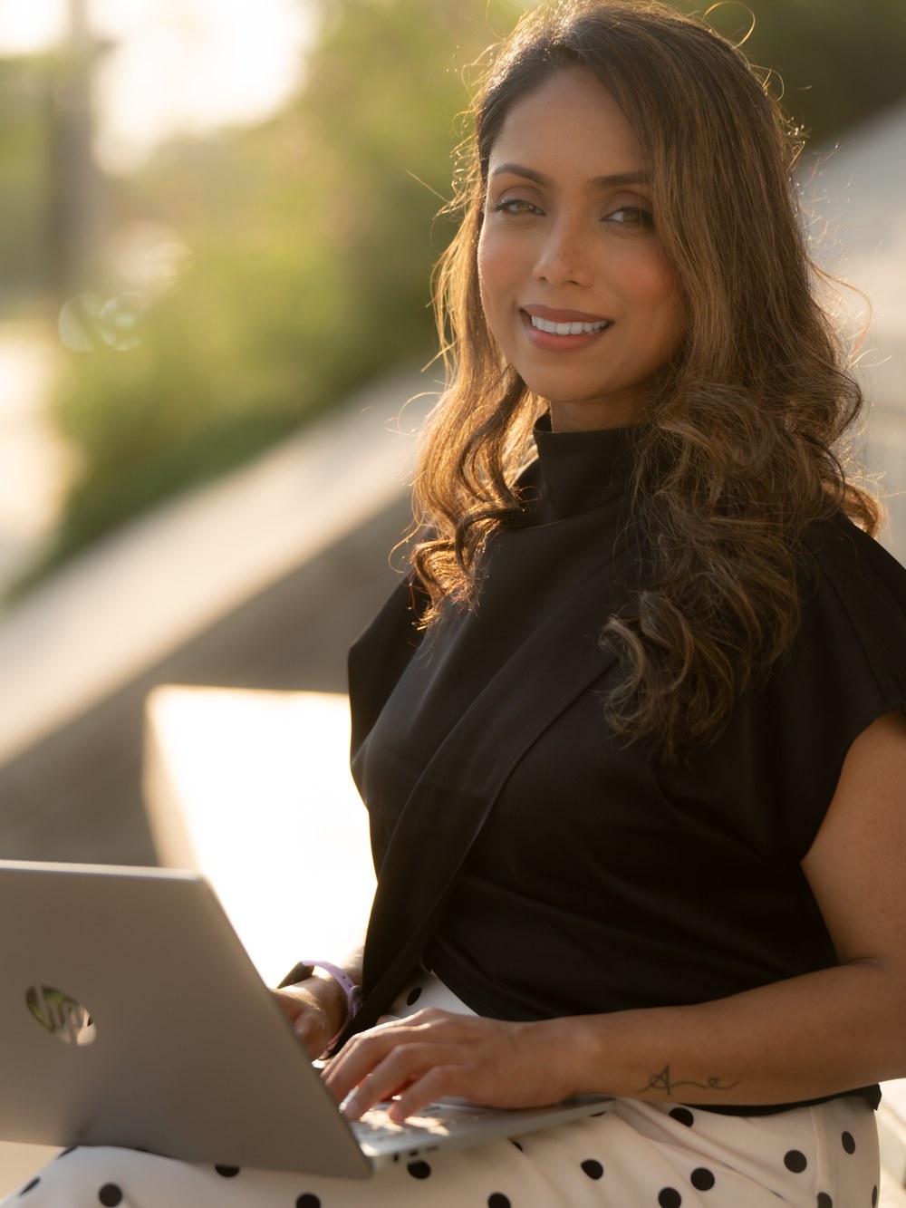 Ferdous seated outdoors in warm evening light, looking up from her laptop with a warm smile